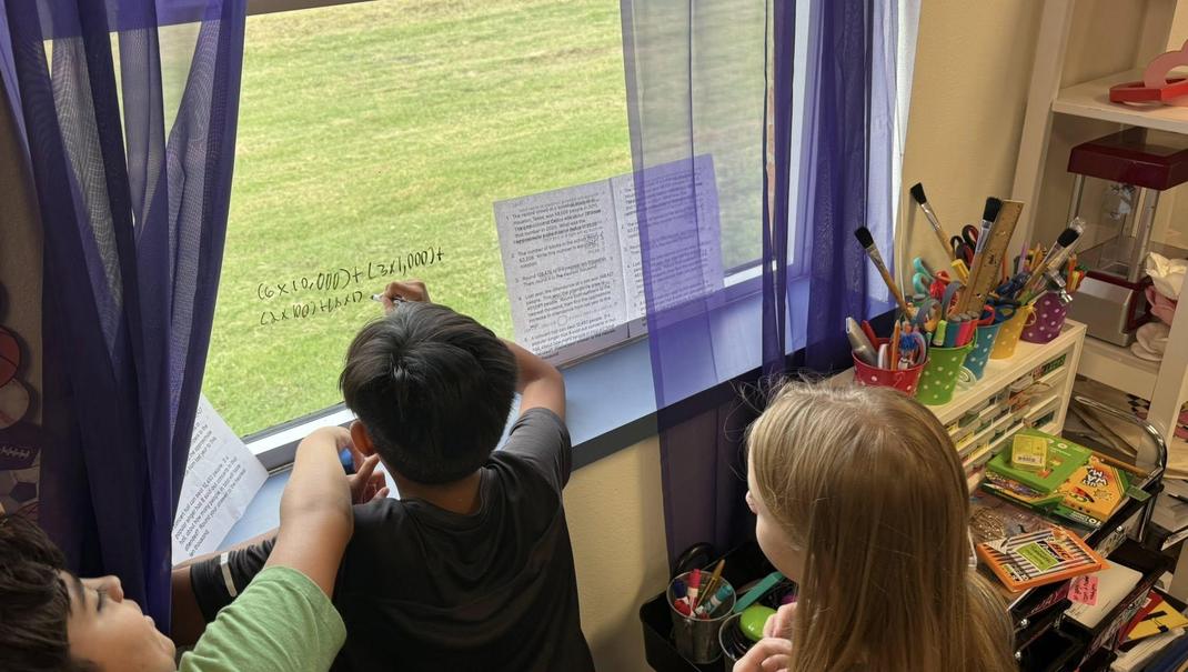 Three children solving math on a window with colorful markers, overlooking a grassy area.