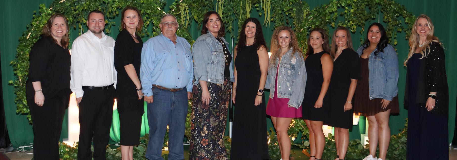 A group of thirteen people posing together in front of green foliage.