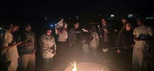 Group of people around a fire at night, some roasting marshmallows.