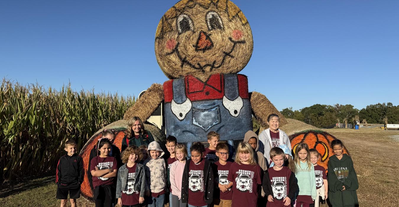 Children in maroon shirts standing in front of a large scarecrow in a cornfield.