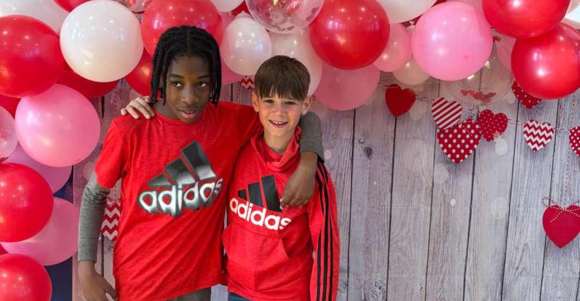 Two children in matching red Adidas shirts pose together in front of a balloon backdrop.