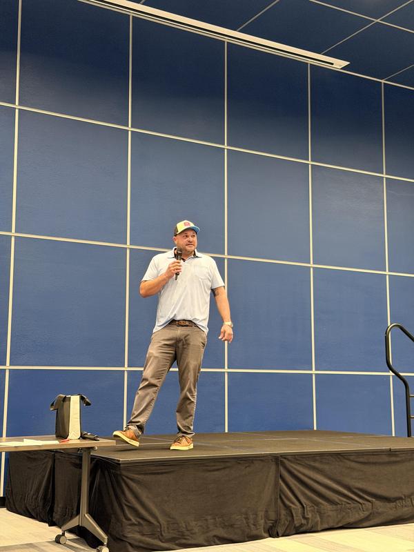 A man speaks on stage in a blue room, holding a microphone and presenting to the audience.