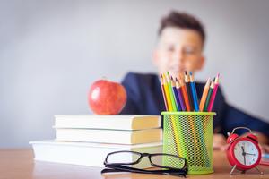 Books, pencils, and an apple on a teachers desk with young boy in the background