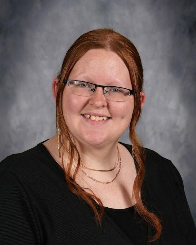 Smiling woman with shoulder-length red hair, glasses, and a black top against a gray backdrop.
