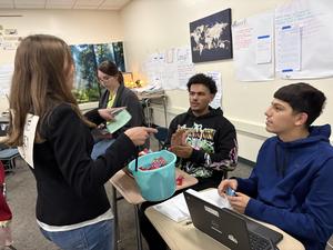 Classroom scene with students exchanging items and interacting at desks with a blue bucket.