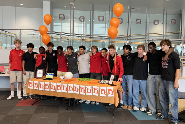 Students stand behind a table with an orange H Logo, soccer ball, and orange balloons