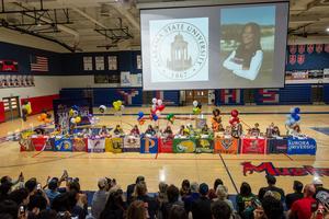 A high school gym is filled with people watching a college signing day event. Eleven students sit at tables, each featuring different college banners and colorful balloons. A large screen behind them displays the Alabama State University logo and a portrait of an individual. The atmosphere is festive and celebratory.