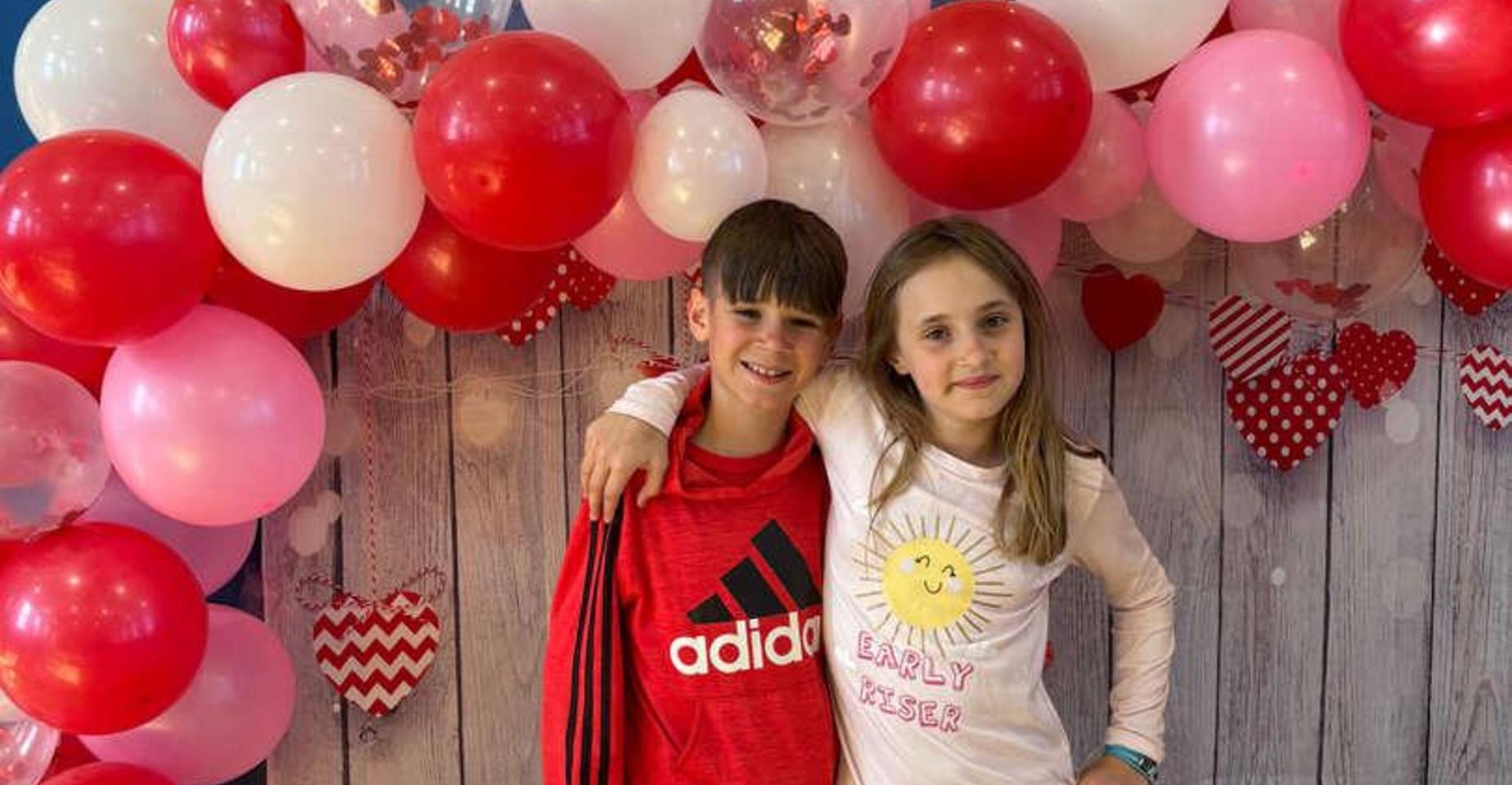 Two children smiling together in front of a colorful balloon display.