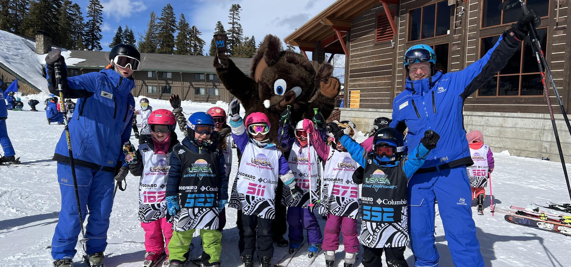 Ski instructors and children celebrating with a large mascot on a snowy ski slope.