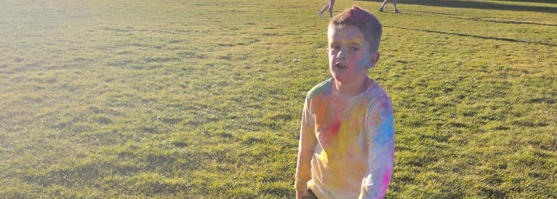 Child with colorful powder on face and shirt, standing on grass in a park.