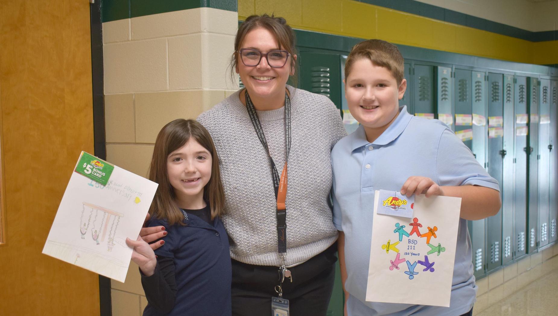 A boy and a girl stand with a teacher holding drawings in a school hallway.