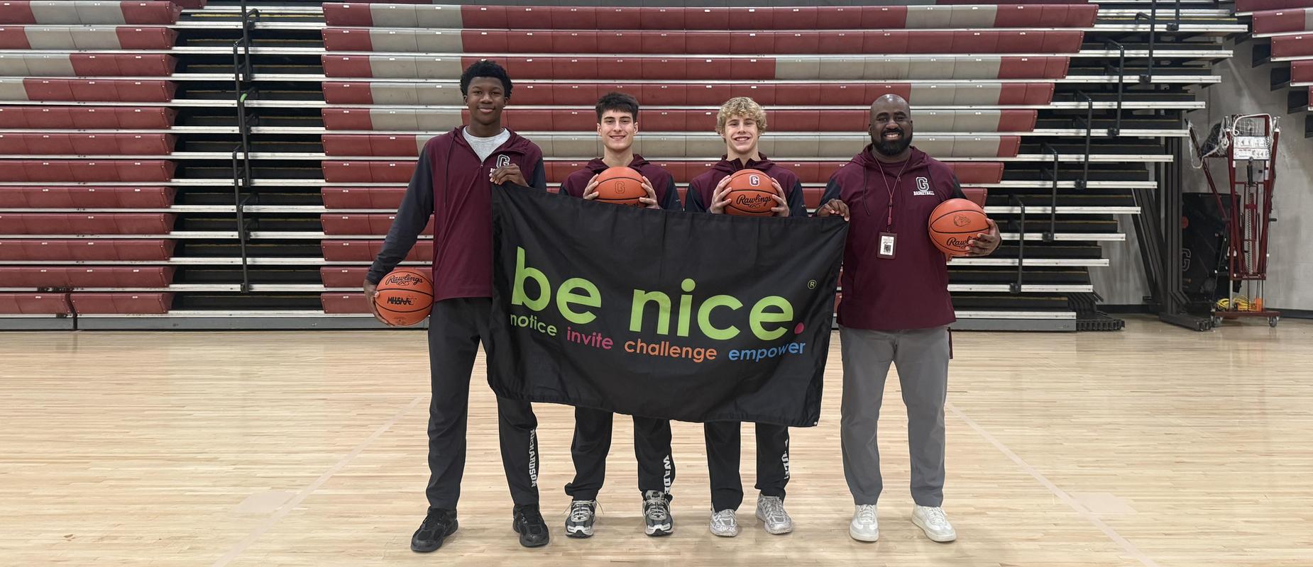 three basketball players and the coach stand in the gym smiling with a be nice banner