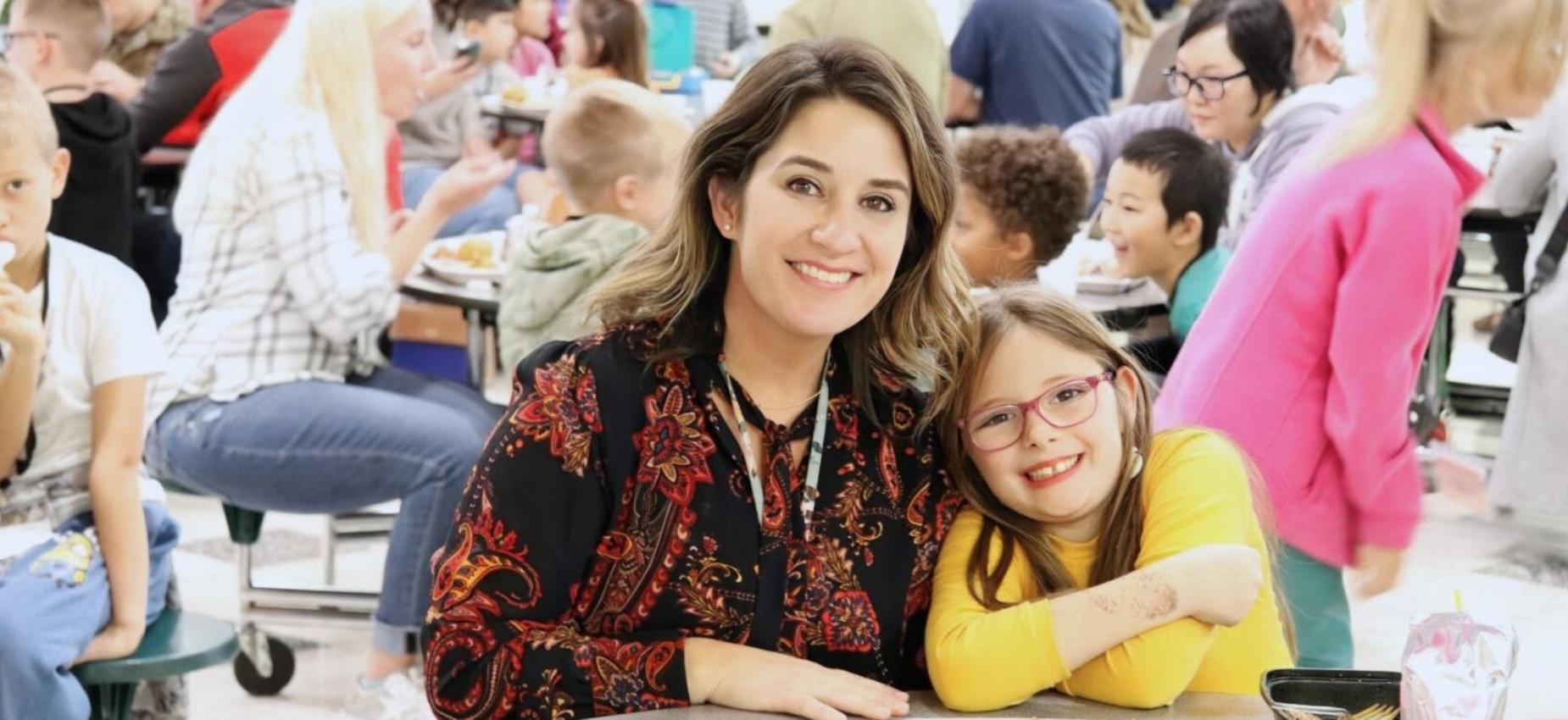 A woman and a girl smiling together in a busy cafeteria.