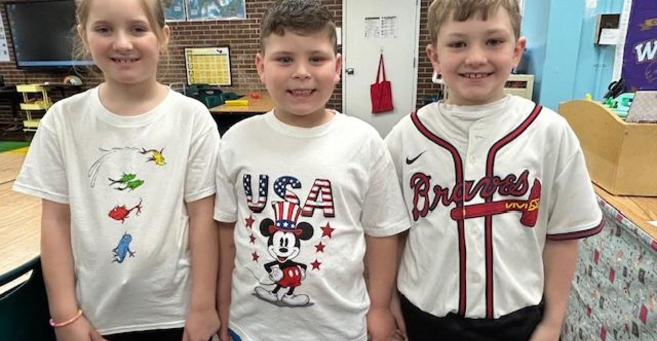 Three children smiling together in a classroom, wearing colorful shirts.