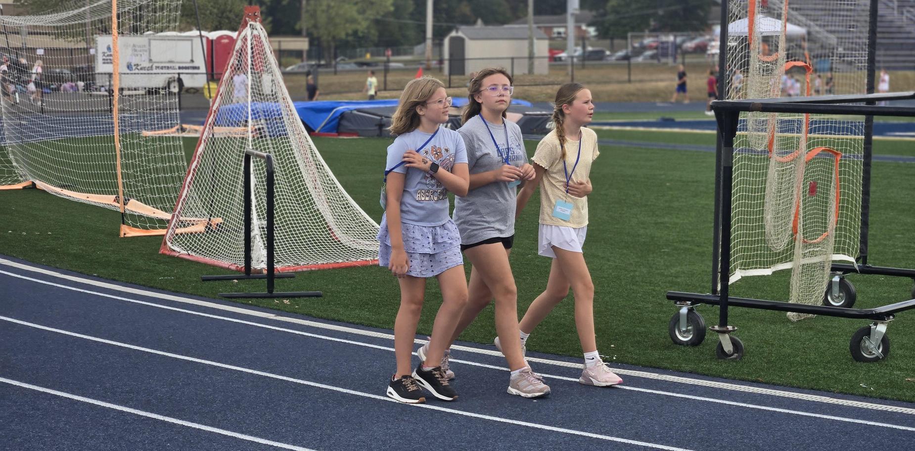 Three girls walk on a running track beside a sports field.