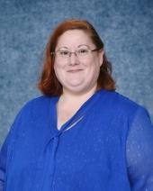 A woman with glasses in a blue shirt, standing against a textured blue backdrop.