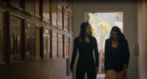 Two women walk side by side down a hallway lined with award plaques. They are engaged in conversation, with natural light streaming in from a doorway ahead, creating a warm and reflective atmosphere.
