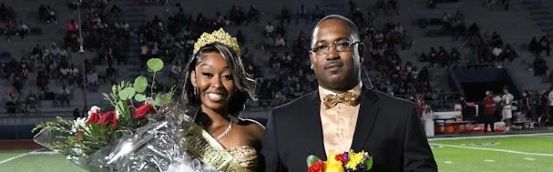 A couple stands on the field holding colorful props and looking at the camera.