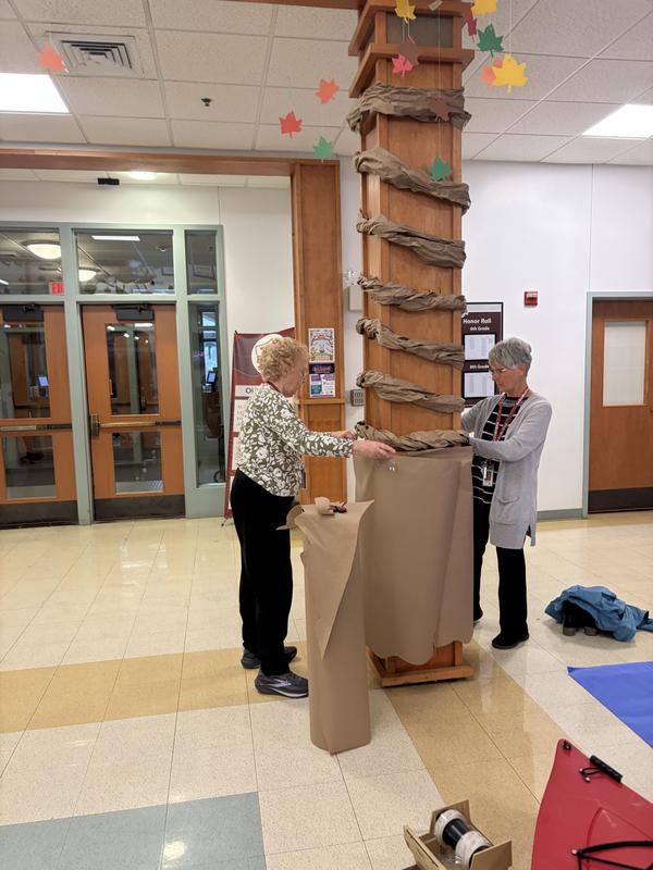 Two individuals wrapping a wooden column with brown paper.