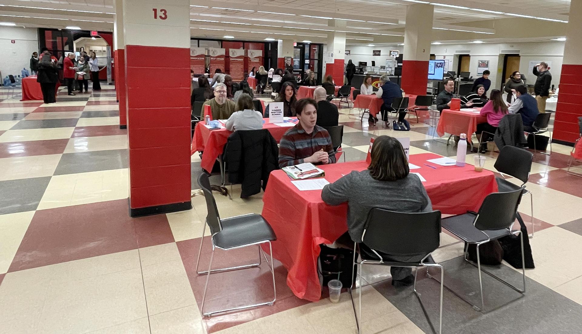 People seated at tables with red tablecloths in a large hall.