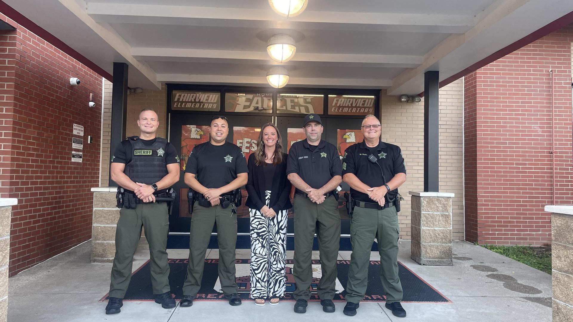 Five law enforcement officers posing with a woman outside a school entrance.