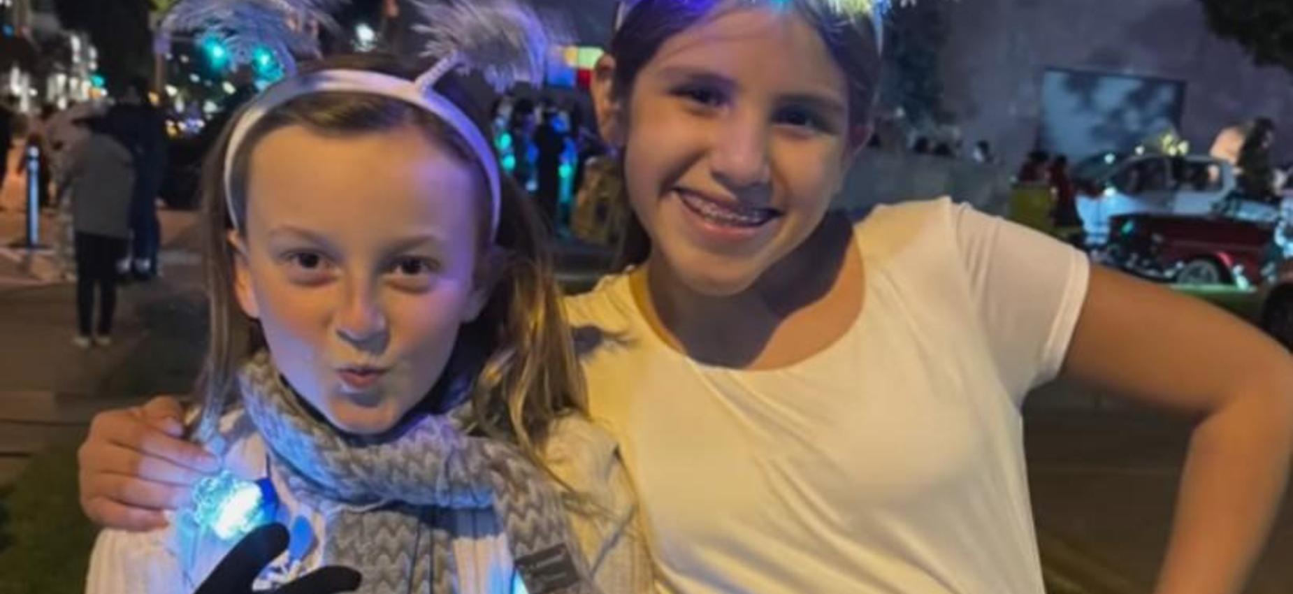 Two smiling girls wearing festive accessories and glowing lights at a night event.