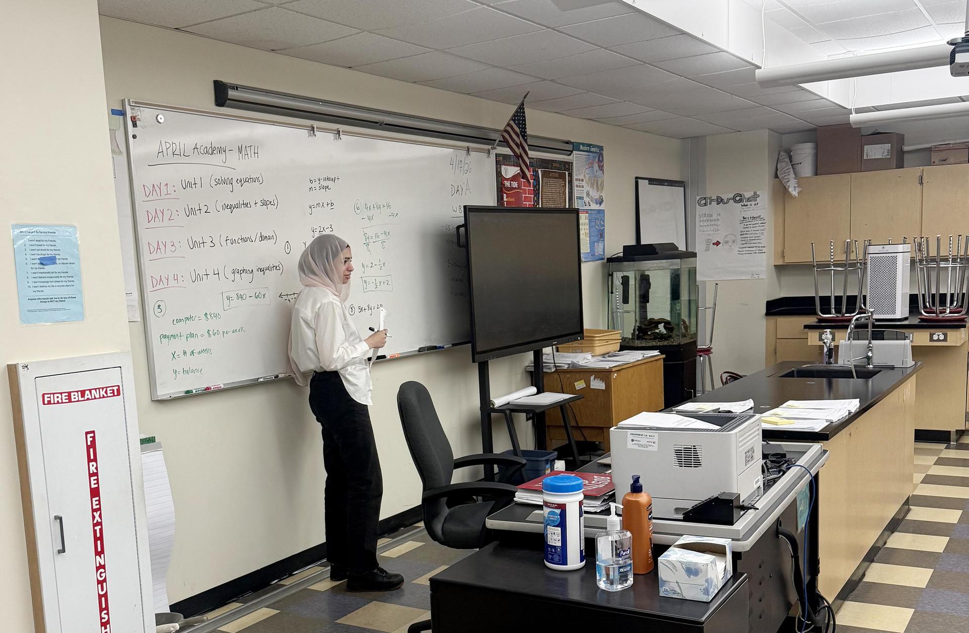 A classroom scene with a teacher writing on a whiteboard and students in the background.
