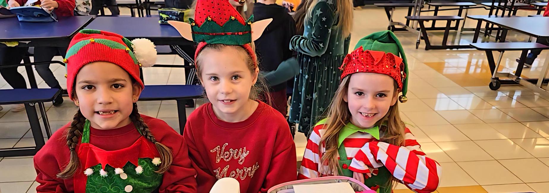 Three smiling students sitting at lunch table with holiday elf hats