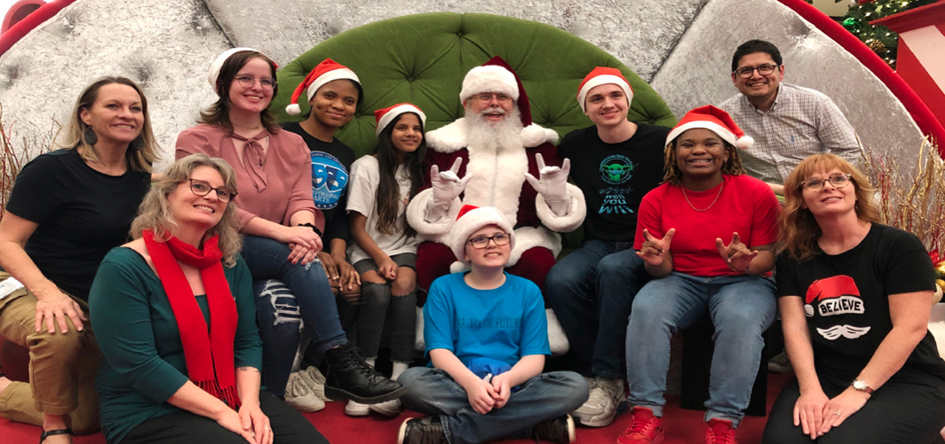 A group photo from the Signing Santa event at Barton Creek Square. Santa sits in a green armchair, surrounded by children and adults, many wearing Santa hats and festive attire. Santa and several attendees are using sign language to spread holiday cheer. The background features holiday decorations, creating a warm and inclusive festive atmosphere.