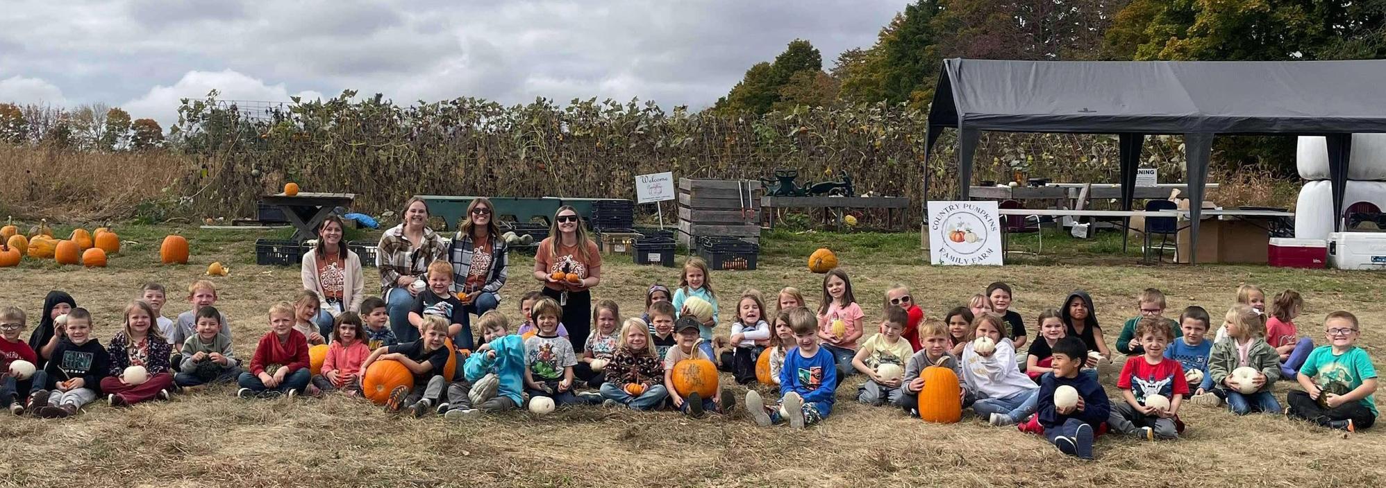 Group of children and adults sitting outdoors among pumpkins and plants.