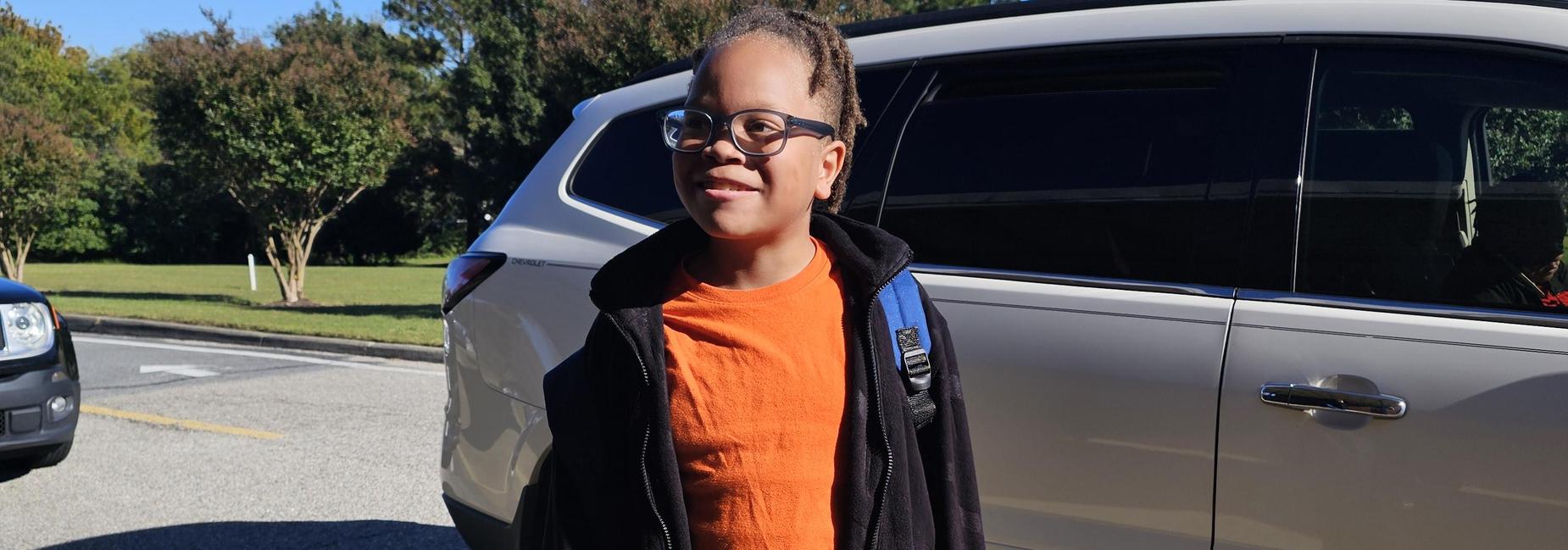 Child in an orange shirt standing near a parked car.