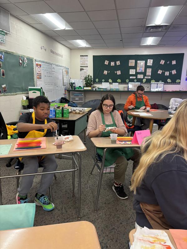 Students engaged in painting at desks in a classroom, focused on their colorful artwork.