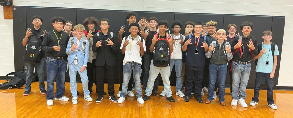 Group of teenage boys posing together in a gymnasium, making hand gestures.