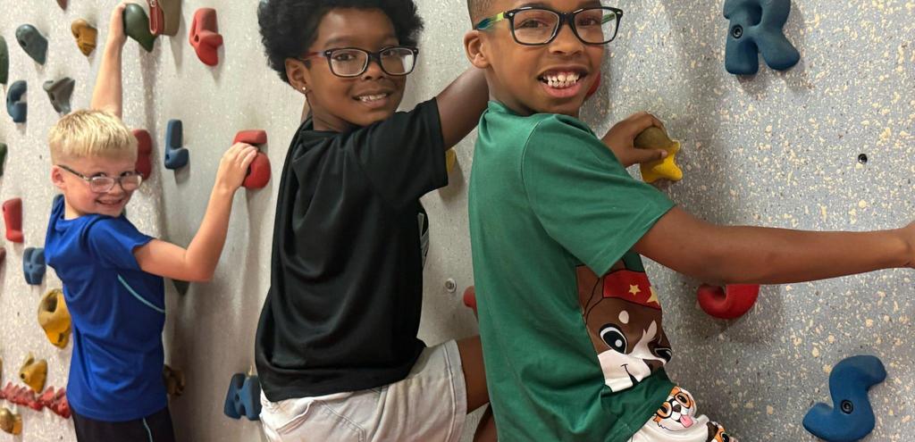 Three children climbing a colorful indoor rock wall, smiling and enjoying the activity.