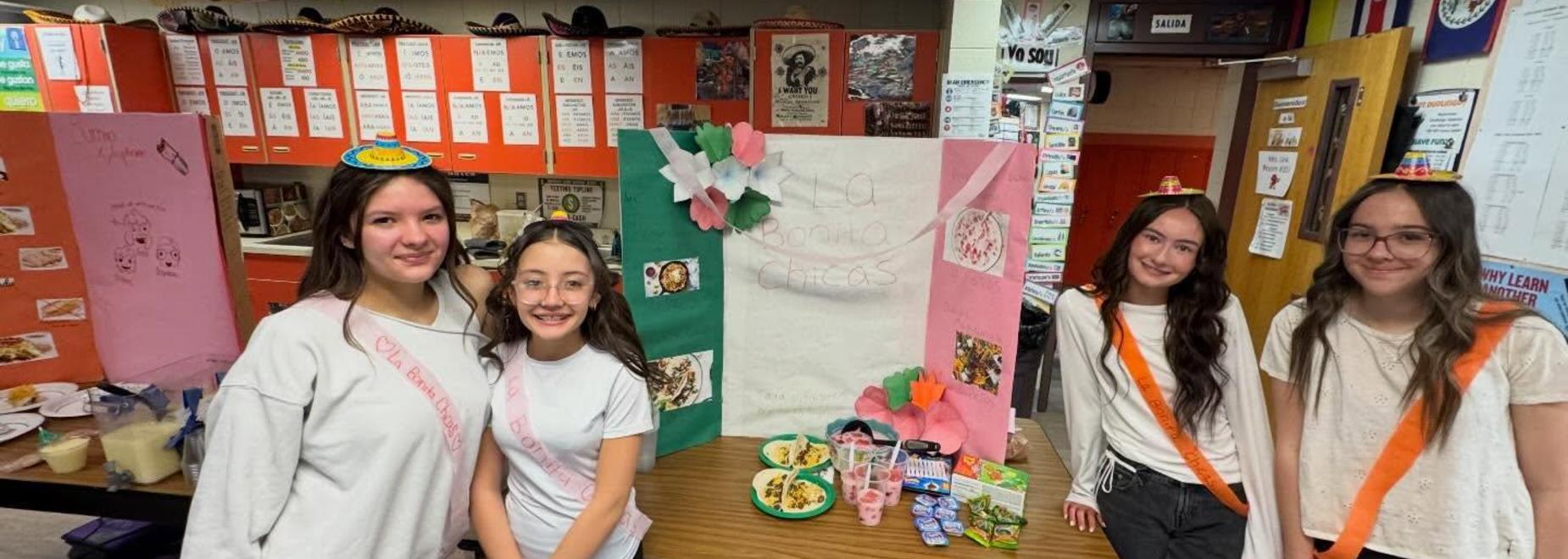 Four students pose with a display featuring Mexican culture and food.