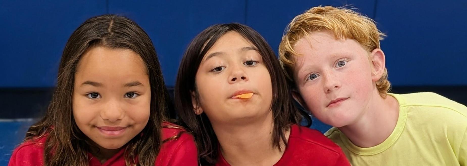 Three children with diverse expressions, one holding a snack, against a blue background.