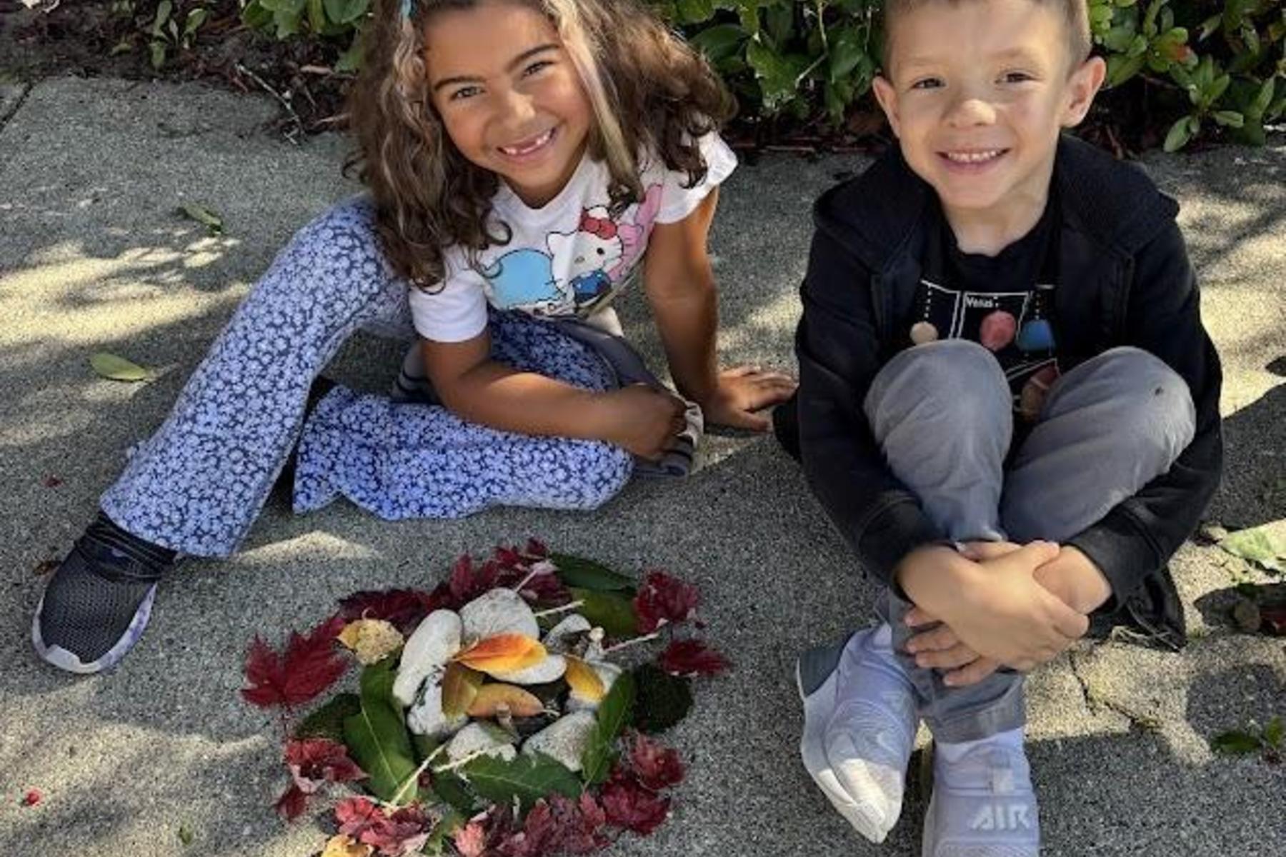 Two kids posing with a nature arrangement on the ground, surrounded by greenery.
