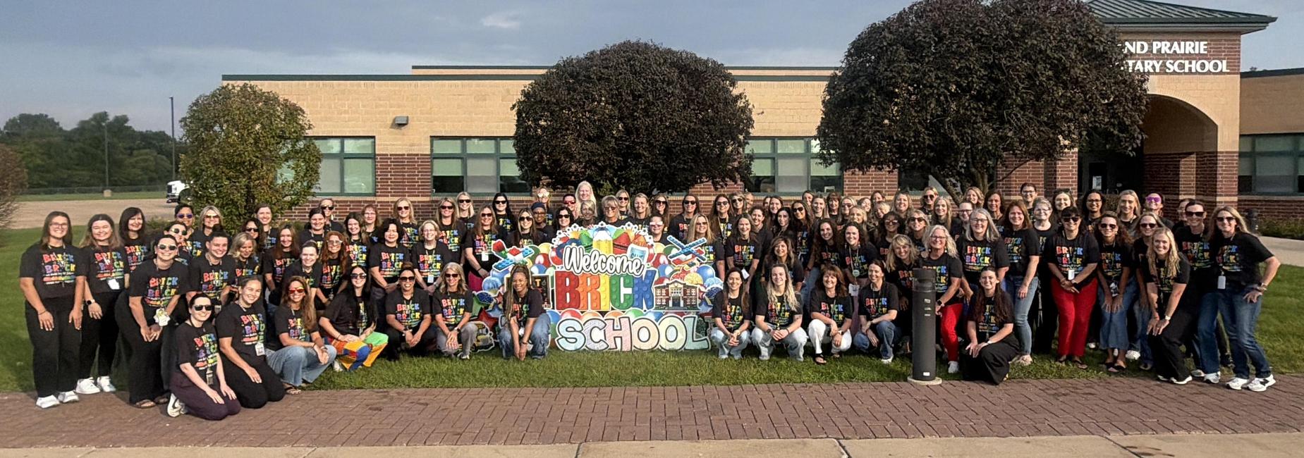 Group of educators posing in front of a school, wearing matching shirts.