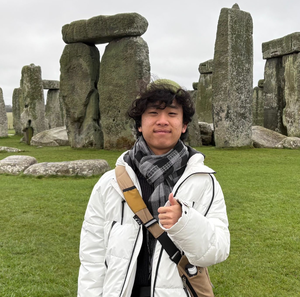 Andrew Kim, a teenage boy with black curly hair wearing a white parka and gray plaid scarf smiling and posing with a thumbs up in front of Stone Henge.