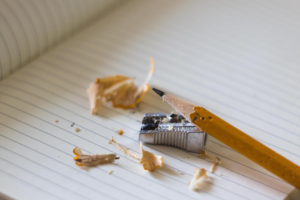 A sharpened yellow pencil resting beside a metal sharpener and pencil shavings on lined paper.