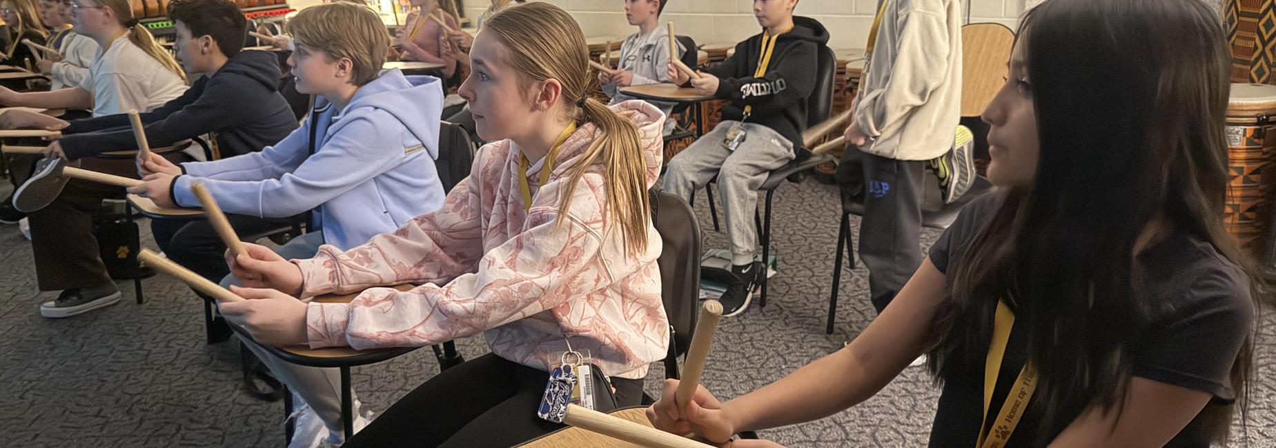 A group of students sitting on stools while playing drums in a classroom setting.