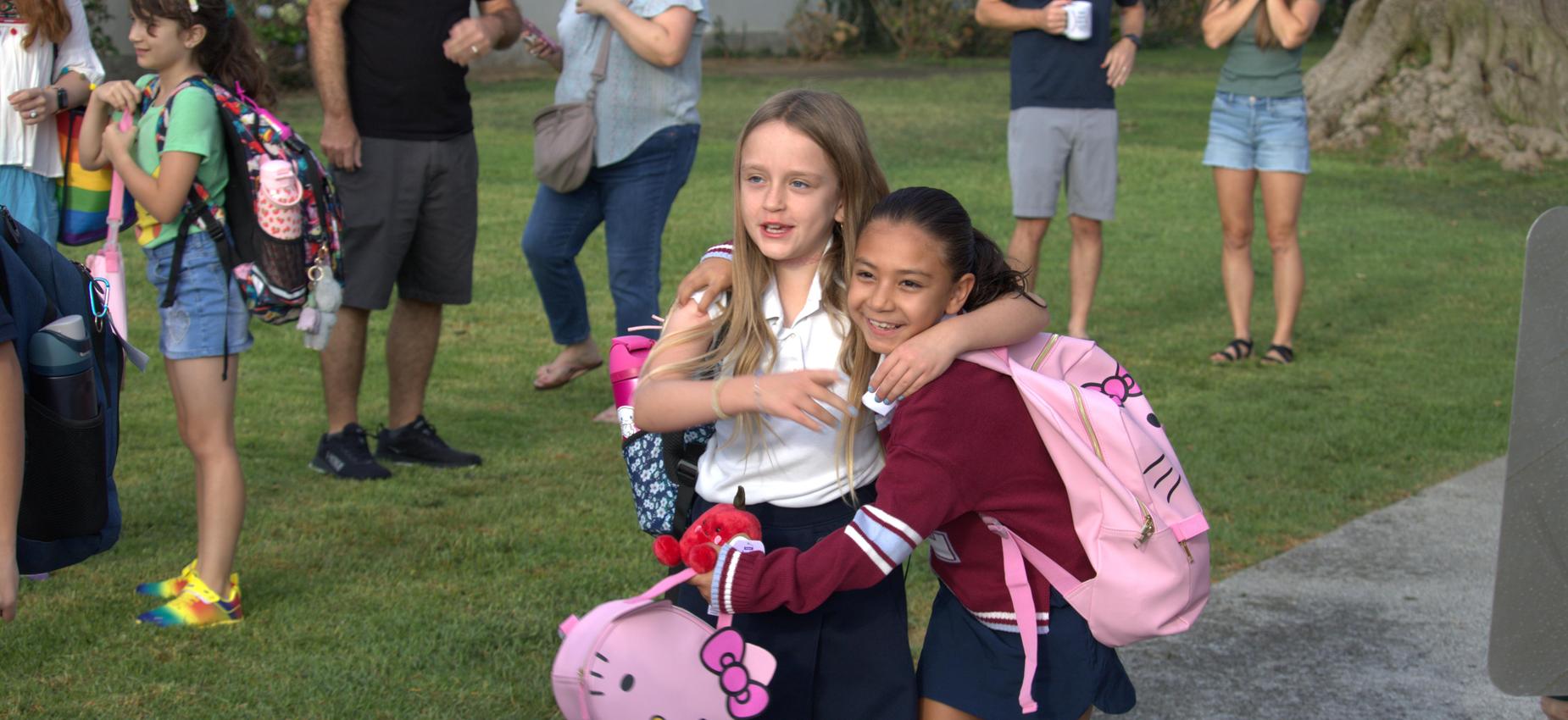 Two girls in school uniforms hug, holding backpacks in a lively outdoor setting.