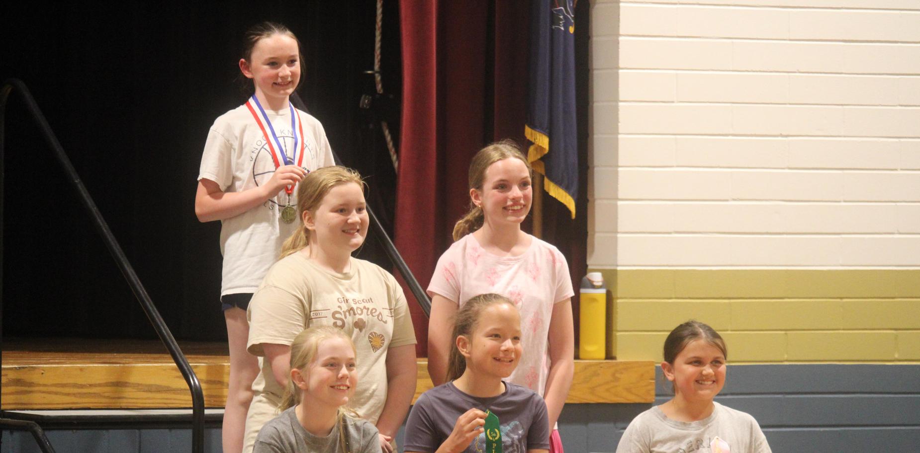 Group of girls seated with medals and ribbons, smiling at an indoor ceremony.