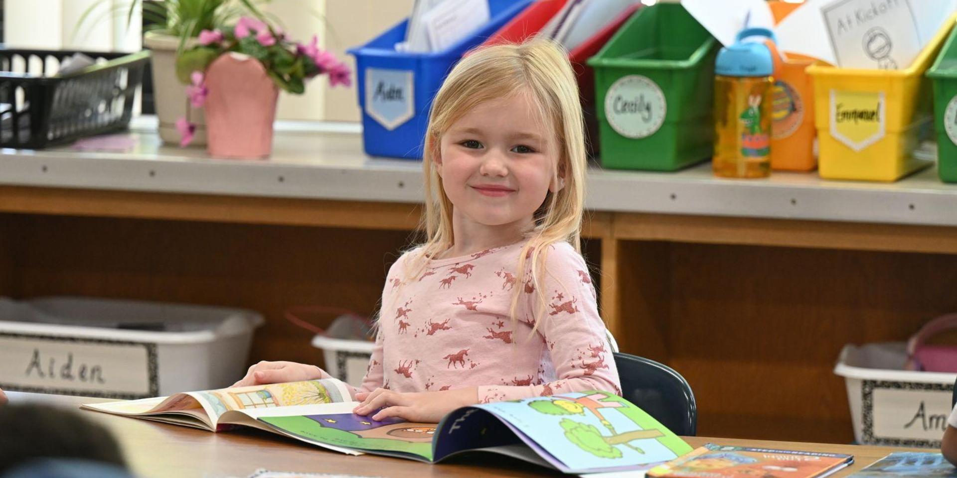 A young girl sitting at a table, smiling while holding a colorful book in a classroom.