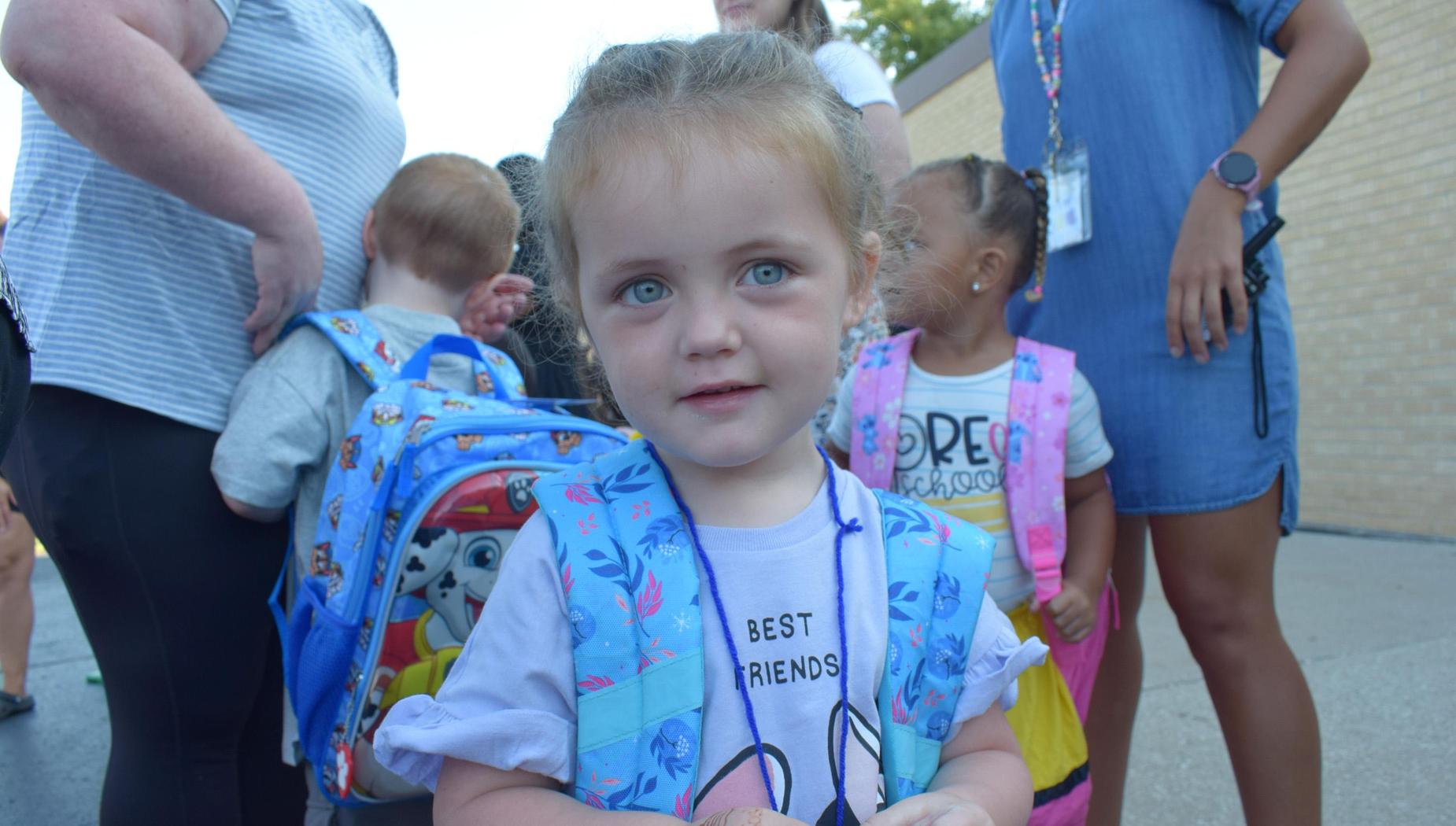 A young girl with a backpack looking at the camera amidst a group of children.