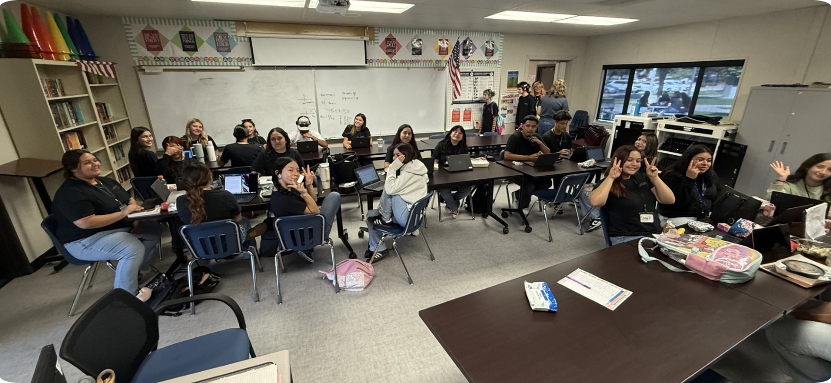 Students in a classroom seated at tables, engaging and smiling at the camera.