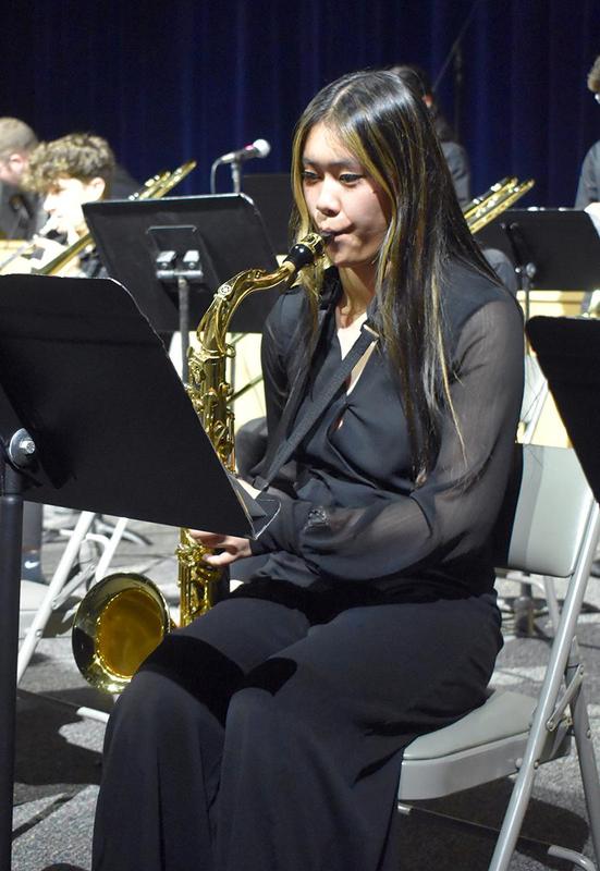 A young woman plays the saxophone in a seated position during a performance.