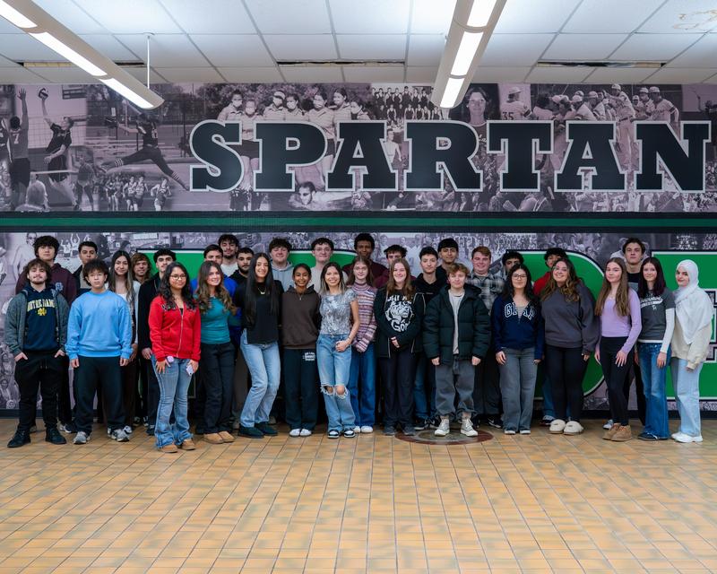 A group of high school students pose together in a school hallway beneath a large “SPARTAN” mural.