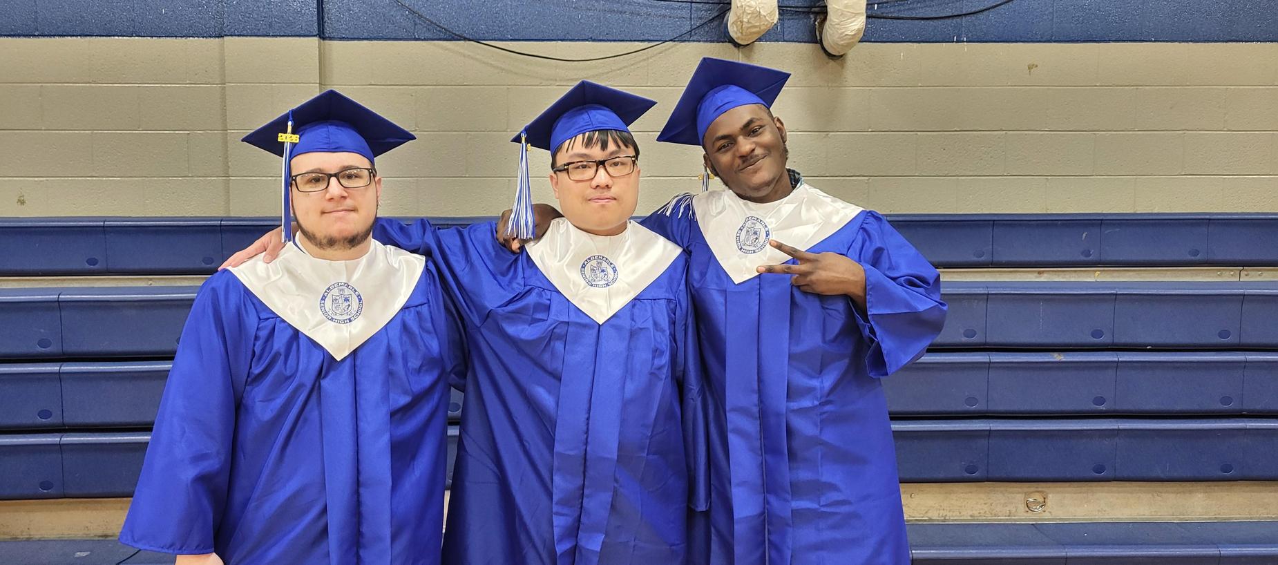 Three graduates in blue caps and gowns pose together in a gymnasium.