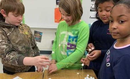 Students pictured gathered around a table using toothpicks and marshmallows to construct square frame